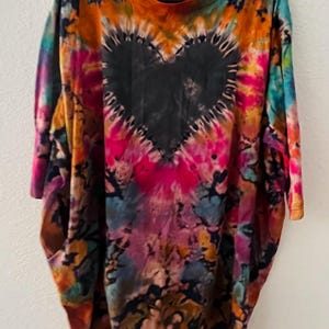 以下が含まれることがあります： 中央に大きな黒いハートのデザインが施されたタイダイTシャツ。オレンジ、ピンク、青、黄色など、鮮やかな色の組み合わせが特徴です。黒いハンガーにかかっています。