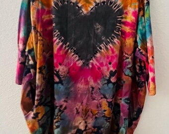 May include: A tie-dye t-shirt with a large black heart design in the center. The shirt features a vibrant mix of colors, including orange, pink, blue, and yellow. The shirt is hanging on a black hanger.