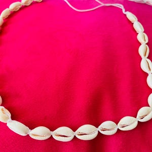 以下が含まれることがあります： 白い紐に通された、クリーム色の貝殻で作られたネックレス。 貝殻は楕円形で、連続したループ状に配置されています。 ネックレスは明るいピンク色の表面に展示されています。