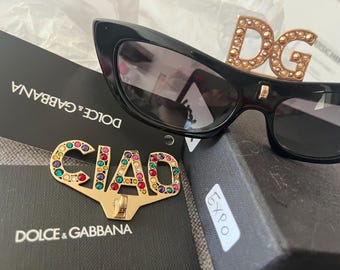 Puede incluir: Gafas de sol negras de ojo de gato con un logotipo DG dorado adornado con pedrería. Un pin "CIAO" dorado, incrustado con gemas de colores, descansa sobre una caja negra. Las gafas de sol y el pin son de la marca Dolce & Gabbana.