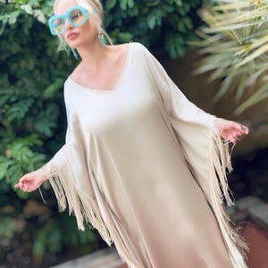 以下が含まれることがあります： Vネック、ワイドスリーブ、両サイドに長いフリンジトリムをあしらった、床まで届く長さのベージュのカフタンドレス。大きめの水色のスクエアフレームサングラスと、ターコイズカラーのぶら下がりイヤリングを合わせています。