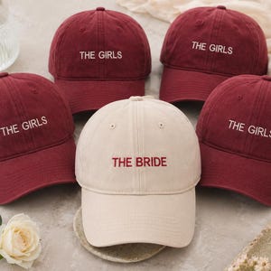 Könnte beinhalten: Fünf Baseballkappen, vier in Burgunderrot und eine in Creme. Die burgunderroten Kappen sind mit "THE GIRLS" in Weiß bestickt, während die cremefarbene Kappe "THE BRIDE" in Burgunderrot trägt. Die Kappen liegen auf einer hellen Oberfläche.