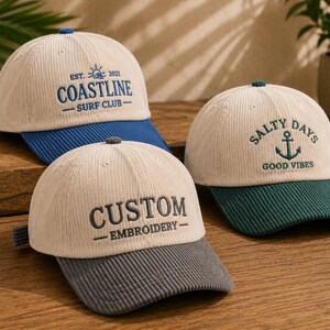 May include: Three corduroy baseball caps with cream-coloured crowns and contrasting brims. One cap features blue embroidery reading COASTLINE SURF CLUB EST. 2021, one has green embroidery reading SALTY DAYS GOOD VIBES with an anchor, and one has grey text CUSTOM EMBROIDERY.