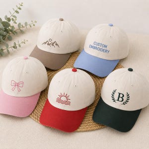 May include: Five baseball caps with off-white crowns and coloured brims. The caps feature embroidered designs: a mountain range with a brown brim, the text CUSTOM EMBROIDERY with a blue brim, a pink bow with a pink brim, a sun over waves with a red brim, and the letter B inside a laurel wreath with a dark green brim.