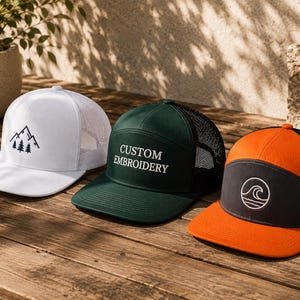 May include: Three trucker caps with mesh backs. The white cap features a black mountain and pine tree graphic. The dark green cap displays the white text CUSTOM EMBROIDERY. The orange and dark grey cap features a white circular wave graphic.