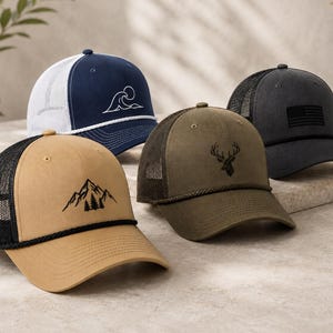 May include: Four trucker caps featuring mesh backs and rope detailing on the brims. The collection includes a tan cap with a mountain graphic, a navy cap with a white wave design, an olive green cap with a deer head emblem, and a charcoal grey flag cap.