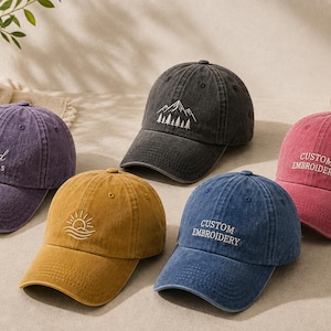 May include: Five baseball caps in purple, mustard yellow, charcoal grey, blue, and red. The purple cap features the text Good Vibes, the grey cap shows a mountain and tree graphic, the yellow cap has a sun and wave design, and the blue and red caps feature the text CUSTOM EMBROIDERY.