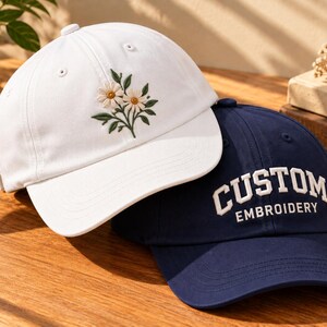 May include: A white baseball cap featuring an embroidered design of two daisies with green leaves and a navy blue baseball cap with the white embroidered text "CUSTOM EMBROIDERY" in a bold, arched font, both with stitched brims and eyelets.