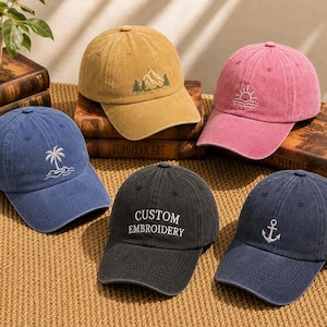 May include: Five baseball caps in blue, tan, pink, black, and dark blue. The blue cap has a palm tree, the tan cap has a mountain and trees, the pink cap has a sun over waves, the black cap has the text CUSTOM EMBROIDERY, and the dark blue cap has an anchor.