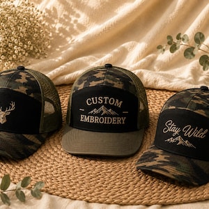May include: Three camouflage trucker hats with mesh backs and solid front panels. The left hat features a gold deer head emblem. The centre hat displays the text CUSTOM EMBROIDERY above a mountain graphic. The right hat shows the text Stay Wild.