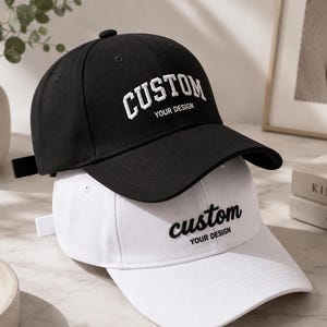 May include: A black baseball cap with white embroidered text reading CUSTOM YOUR DESIGN and a white baseball cap with black embroidered text reading custom YOUR DESIGN, both featuring a curved brim and a button on the top of the crown.