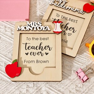 May include: Two wooden picture frames with the text "To the best Teacher ever" and "Mrs Montoya" and "Balderrama". Each frame has a red apple accent. Other items include a pink sticky note pad, pens, and yellow sunglasses.