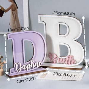 May include: Two wooden letter decorations, a lavender "D" and a white "B", each with a name. The "D" is 18cm (7.09in) tall and 20cm (7.87in) wide, with the name "Daphne". The "B" is 25cm (9.84in) tall and 23cm (9.06in) wide, with the name "Brielle".
