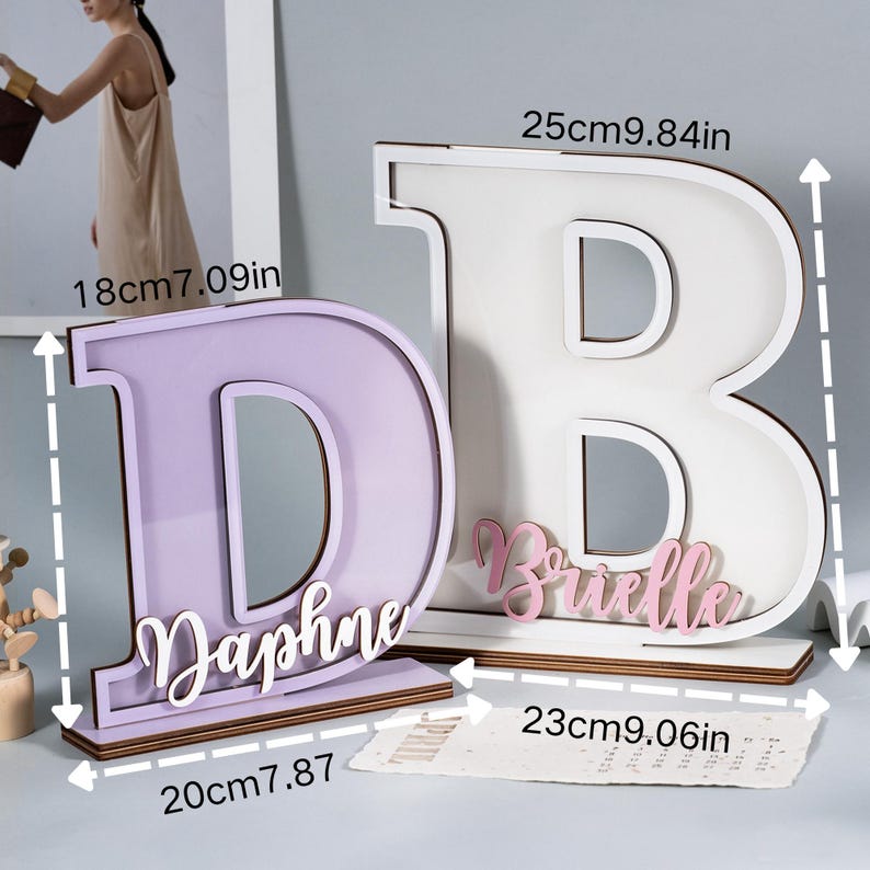 May include: Two wooden letter decorations, a lavender "D" and a white "B", each with a name. The "D" is 18cm (7.09in) tall and 20cm (7.87in) wide, with the name "Daphne". The "B" is 25cm (9.84in) tall and 23cm (9.06in) wide, with the name "Brielle".