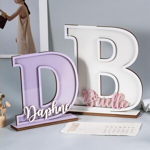 May include: Two wooden letter decorations, a lavender "D" with "Daphne" and a white "B" with "Brielle" in pink script. Both letters are on a wooden base, perfect for home decor or personalized gifts.