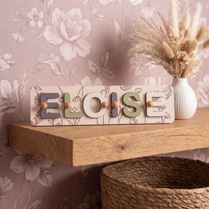 May include: A rectangular wooden name sign featuring the name ELOISE in raised, multi-colored block letters. The sign has a light background with a delicate brown floral line art pattern and small wooden pegs attached to each letter.