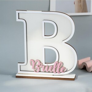 May include: A large, white wooden letter "B" with a white border and a wooden base. The name "Brielle" is written in pink script across the bottom of the letter. The background is a light gray.