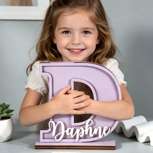 May include: A lavender wooden letter "D" with the name "Daphne" in white script. The letter is a decorative item, possibly a room decoration or a personalized gift. The item is sitting on a brown base.