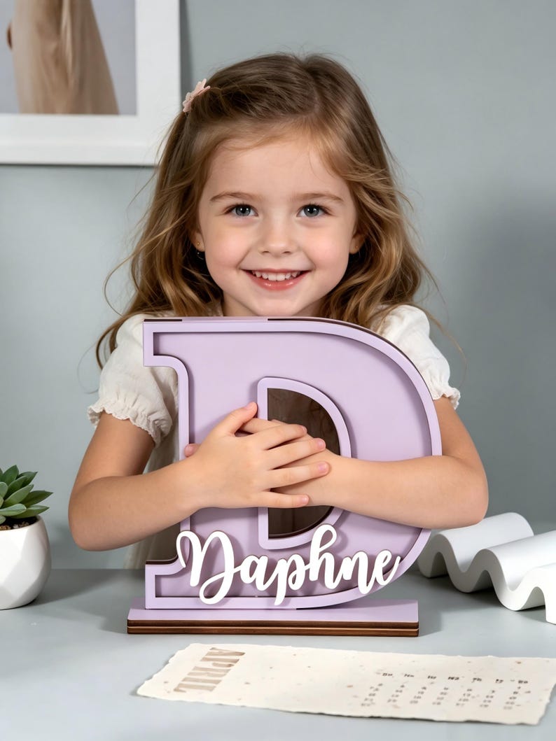 May include: A lavender wooden letter "D" with the name "Daphne" in white script. The letter is a decorative item, possibly a room decoration or a personalized gift. The item is sitting on a brown base.