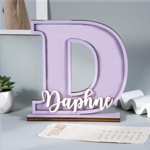May include: A lavender-colored wooden letter "D" with a mirror insert, and the name "Daphne" in white script. The letter sits on a wooden base, suitable for a desk or shelf decoration.