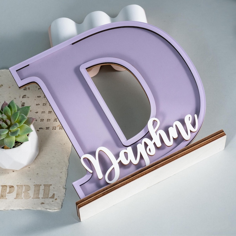 May include: A lavender wooden letter "D" with the name "Daphne" in white script. The letter is mounted on a wooden base with a white front. A small succulent in a white pot and a calendar are in the background.