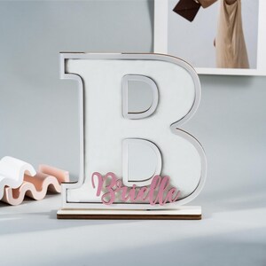 May include: A large white wooden letter "B" with a silver outline and a pink nameplate that reads "Brielle". The letter sits on a wooden base. Decorative item for a nursery or child's room.