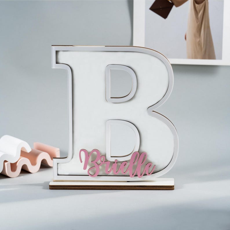 May include: A large white wooden letter "B" with a silver outline and a pink nameplate that reads "Brielle". The letter sits on a wooden base. Decorative item for a nursery or child's room.