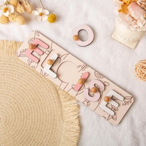 May include: A wooden name puzzle spelling "ELISE" with pink and white letters and wooden pegs. The puzzle board is light beige with a floral design. The letter "O" is separate. The puzzle is on a white, textured surface.
