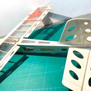 May include: A partially assembled model airplane, featuring a wooden frame with transparent covering on the wings. The design includes a red cross detail and oval cutouts on the tail section. The model is set against a green cutting mat.