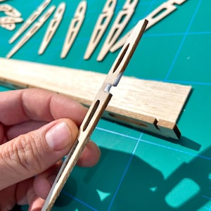 May include: Close-up of wooden model airplane parts. A hand holds a thin, light-colored wooden piece with a slot and a white connector. Other wooden pieces, including ribs and a longer spar, are visible on a green cutting mat.