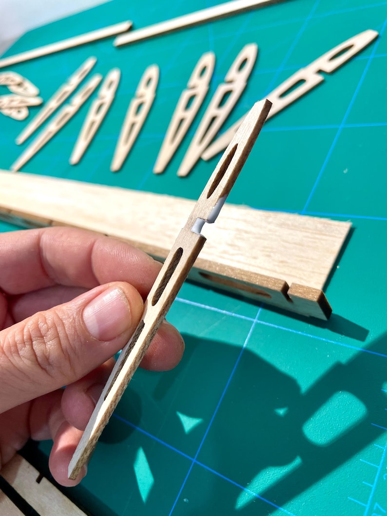 May include: Close-up of wooden model airplane parts. A hand holds a thin, light-colored wooden piece with a slot and a white connector. Other wooden pieces, including ribs and a longer spar, are visible on a green cutting mat.