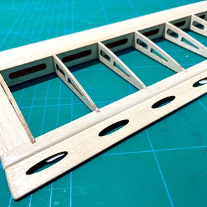 May include: Close-up of a wooden model airplane wing frame. The frame is constructed of light-colored wood with a series of rectangular and triangular supports. The wing has several oval-shaped cutouts along its edge. The background is a green cutting mat.