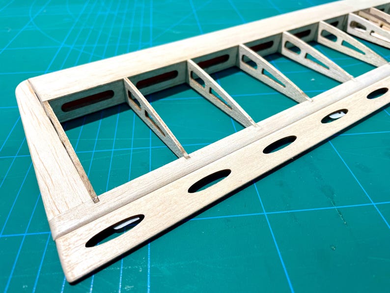 May include: Close-up of a wooden model airplane wing frame. The frame is constructed of light-colored wood with a series of rectangular and triangular supports. The wing has several oval-shaped cutouts along its edge. The background is a green cutting mat.
