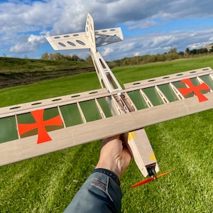 May include: A wooden model airplane with a light brown frame, green panels, and red cross markings on the wings. The propeller is orange. The plane is held against a backdrop of green grass and a cloudy sky.