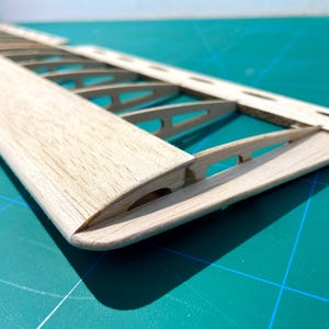 May include: Close-up of a wooden model airplane wing. The wing is constructed with a light-colored wood, featuring a curved leading edge and internal rib supports. The wing is set against a teal cutting mat.