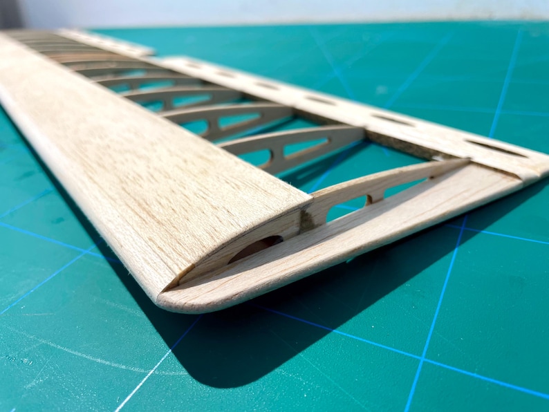 May include: Close-up of a wooden model airplane wing. The wing is constructed with a light-colored wood, featuring a curved leading edge and internal rib supports. The wing is set against a teal cutting mat.