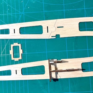 May include: Two wooden airplane wing components with rectangular and square cutouts, along with smaller wooden pieces, are arranged on a green cutting mat. The components are light brown and appear to be part of a model airplane kit.