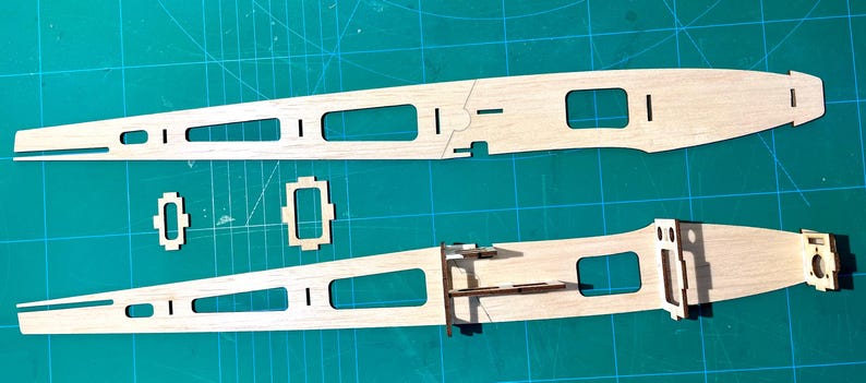 May include: Two wooden airplane wing components with rectangular and square cutouts, along with smaller wooden pieces, are arranged on a green cutting mat. The components are light brown and appear to be part of a model airplane kit.