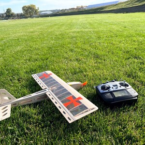 May include: A model airplane with red cross markings on the wings and a remote control sit on a grassy field. The airplane is white and gray with an orange propeller. The remote control is black with a digital display.