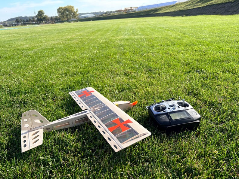 May include: A model airplane with red cross markings on the wings and a remote control sit on a grassy field. The airplane is white and gray with an orange propeller. The remote control is black with a digital display.