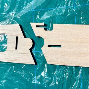 May include: Two unfinished wooden pieces with cut-out shapes, likely for a model or craft project. The pieces are light brown and have rectangular and curved cutouts. They are positioned against a green cutting mat.