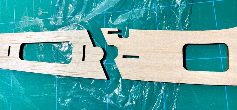 May include: Two unfinished wooden pieces with cut-out shapes, likely for a model or craft project. The pieces are light brown and have rectangular and curved cutouts. They are positioned against a green cutting mat.
