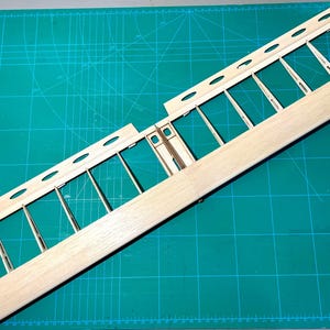 May include: A wooden model airplane wing frame, featuring a light-colored wood construction. The frame has a series of vertical supports and oval-shaped cutouts along the top edge. The wing is positioned on a green cutting mat.