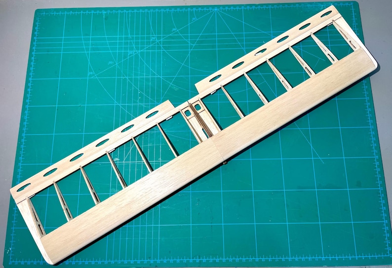 May include: A wooden model airplane wing frame, featuring a light-colored wood construction. The frame has a series of vertical supports and oval-shaped cutouts along the top edge. The wing is positioned on a green cutting mat.