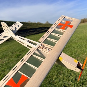 May include: A model airplane with a wooden frame, featuring a red cross on the wing and an orange propeller. The plane has a white tail with black oval cutouts and is set against a green field and blue sky.