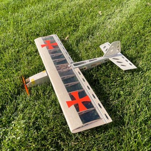 May include: A model airplane with a wooden frame and a red propeller rests on green grass. The wings feature red crosses, and the tail has a unique design. The plane is a vintage-style toy.