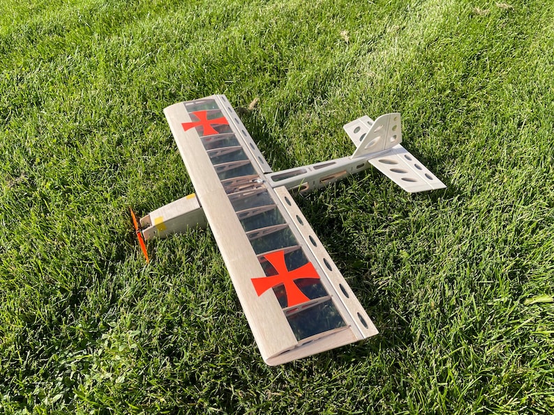 May include: A model airplane with a wooden frame and a red propeller rests on green grass. The wings feature red crosses, and the tail has a unique design. The plane is a vintage-style toy.