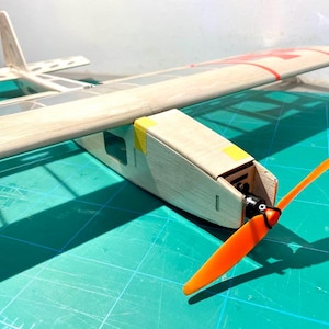 May include: A model airplane with a wooden fuselage and wings. The wings have red accents and the plane has an orange propeller. The model is on a green cutting mat.