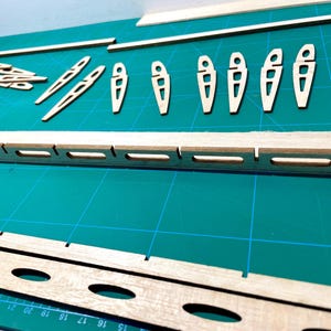 May include: A collection of wooden model airplane parts, including wing ribs, spars, and fuselage components, arranged on a green cutting mat. The parts are made of light-colored wood and are precisely cut with various shapes and openings.