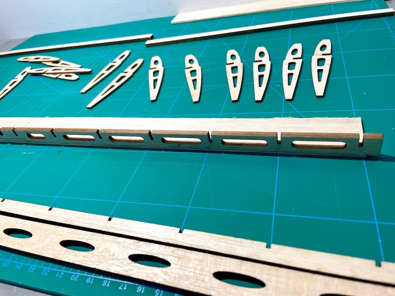 May include: A collection of wooden model airplane parts, including wing ribs, spars, and fuselage components, arranged on a green cutting mat. The parts are made of light-colored wood and are precisely cut with various shapes and openings.
